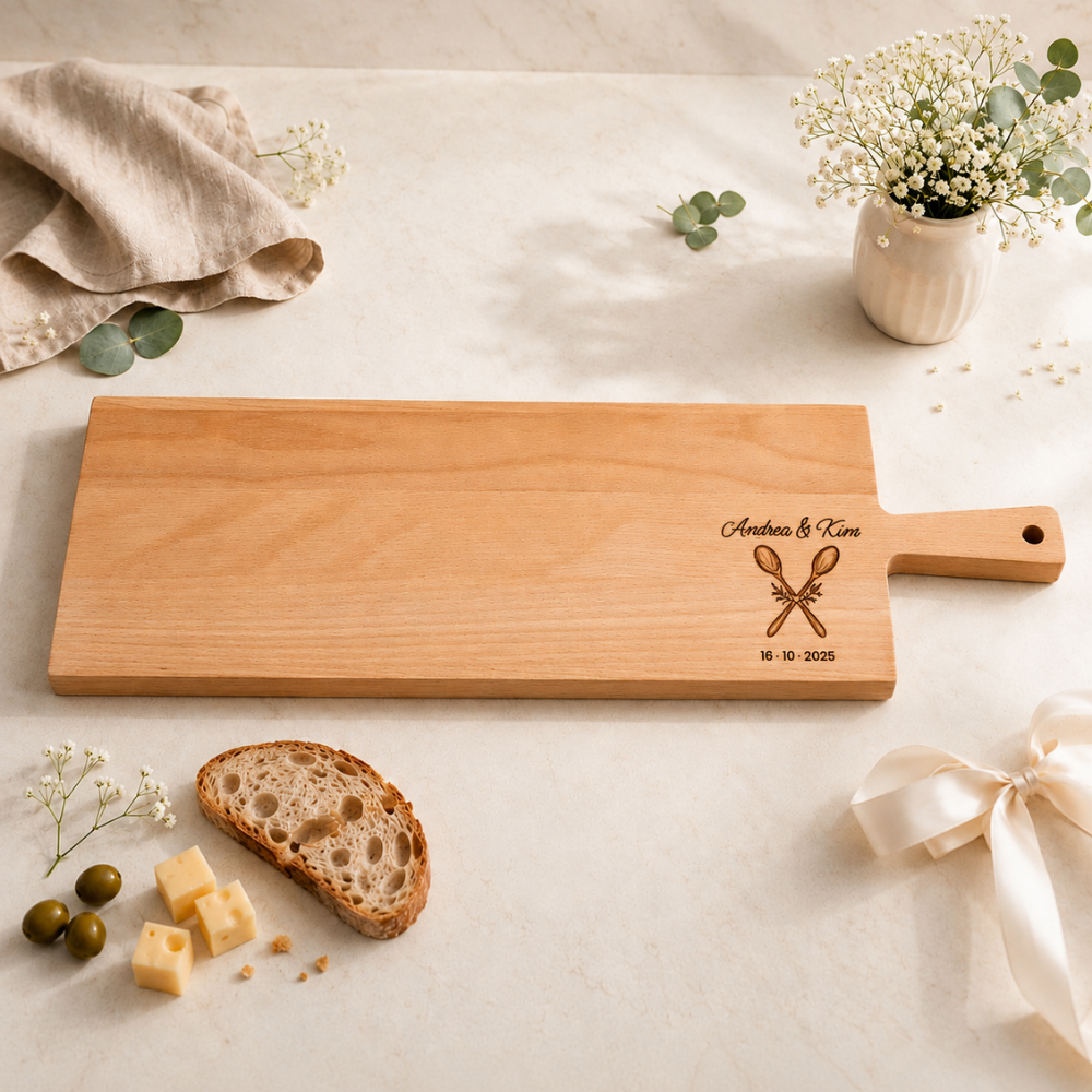 Servierbrett und Apérobrett aus Buchenholz mit Griff, personalisiert mit Hochzeitsgravur: Namen des Brautpaars, zwei gekreuzte Kochlöffel und Hochzeitsdatum, daneben Brot, Käse und Oliven